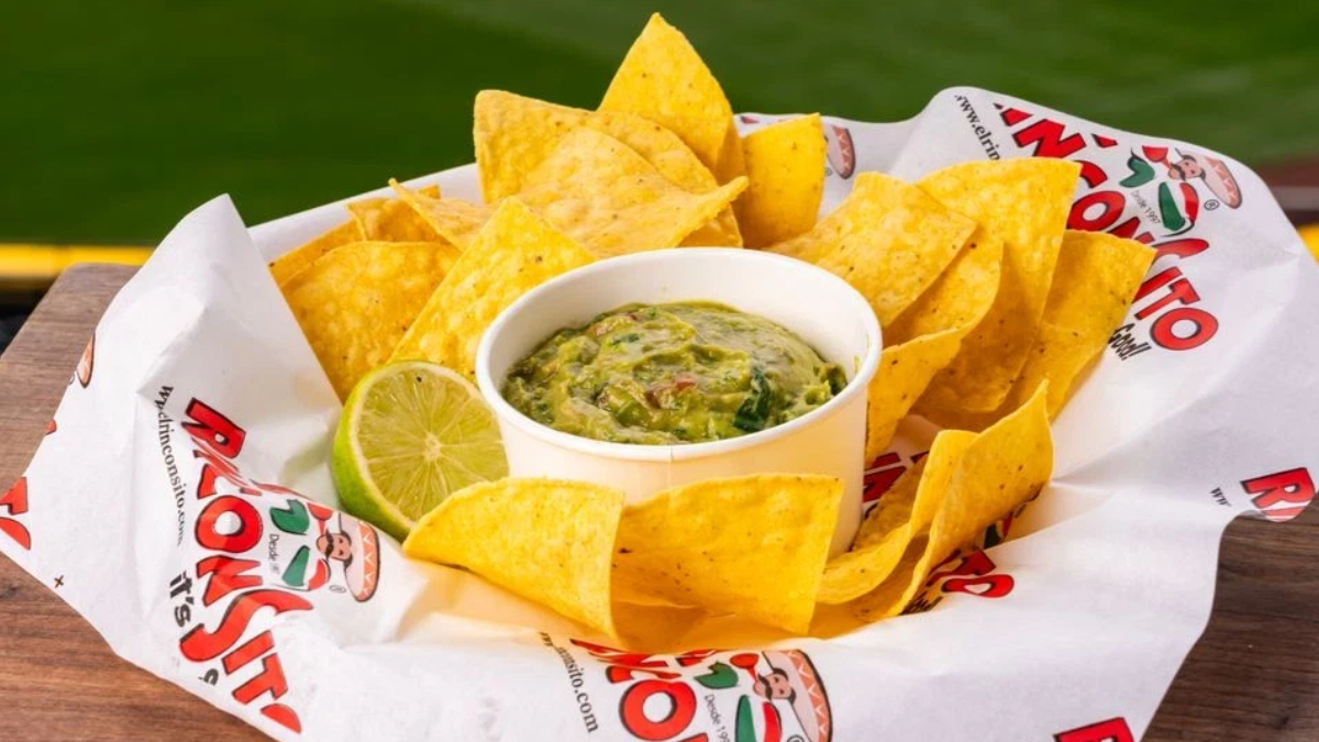 Tortilla chips arranged around a white cup of chunky guacamole with a lime wedge, served on a parchment liner outdoors on a wooden table.