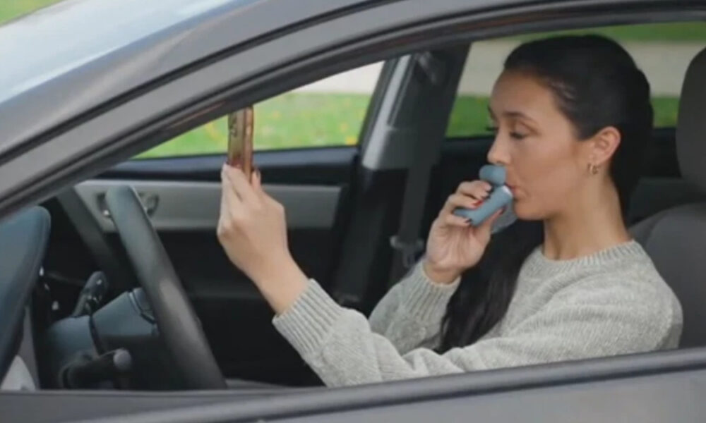 Woman sits in the driver's seat of a car and uses a blue inhaler held to her mouth. She holds a small rectangular object in the other hand, looking forward.