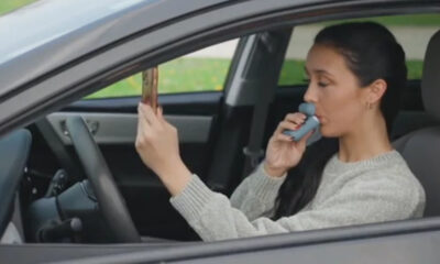 Woman sits in the driver's seat of a car and uses a blue inhaler held to her mouth. She holds a small rectangular object in the other hand, looking forward.