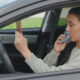 Woman sits in the driver's seat of a car and uses a blue inhaler held to her mouth. She holds a small rectangular object in the other hand, looking forward.