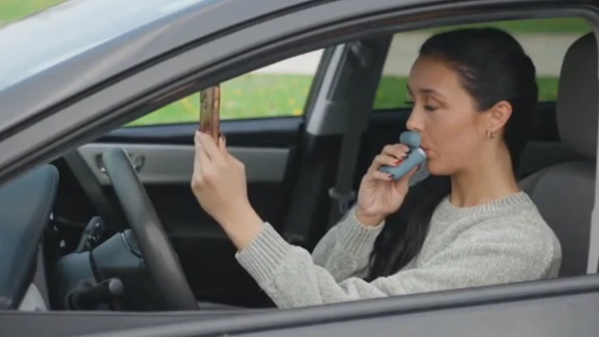 Woman sits in the driver's seat of a car and uses a blue inhaler held to her mouth. She holds a small rectangular object in the other hand, looking forward.