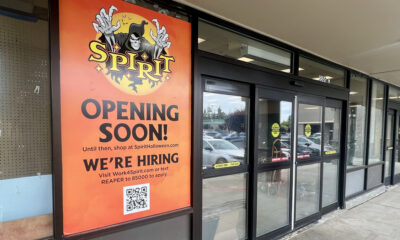 Storefront with a tall orange Spirit Halloween poster reading 'OPENING SOON!' and 'WE'RE HIRING,' including a QR code at the bottom.