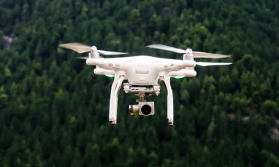 White quadcopter drone with a mounted camera hovering above a dense forest.”,