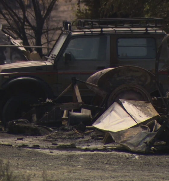 Burned vehicles and scattered debris in a scorched lot after a blaze; a damaged SUV sits among charred wreckage and rusty metal remains.
