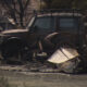Burned vehicles and scattered debris in a scorched lot after a blaze; a damaged SUV sits among charred wreckage and rusty metal remains.