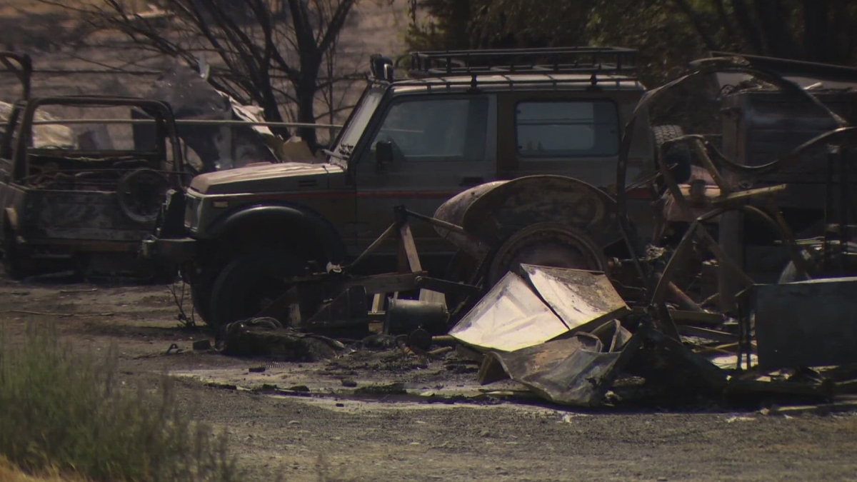 Burned vehicles and scattered debris in a scorched lot after a blaze; a damaged SUV sits among charred wreckage and rusty metal remains.