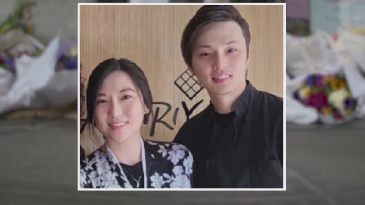 Two young adults, a woman and a man, smiling for a photo side by side indoors with a wooden backdrop behind them.