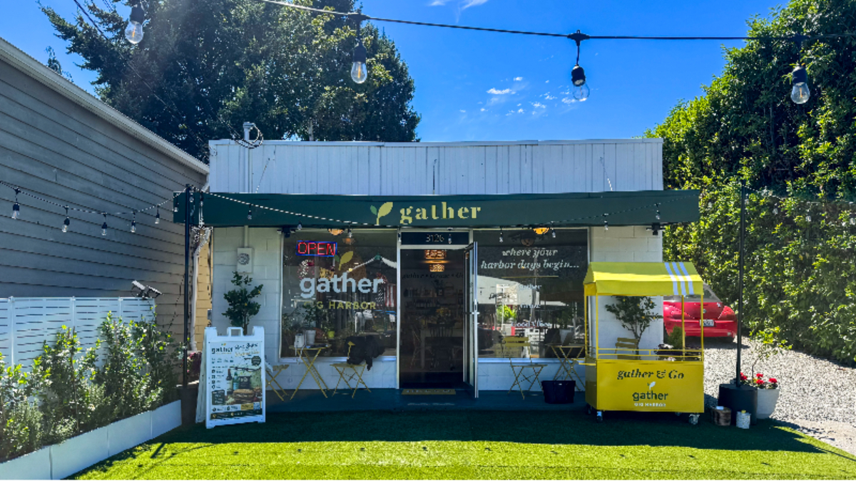 Small storefront with a green awning reading 'gather' and an Open sign; outdoor seating and a yellow 'gather & go' cart on the right, string lights above.