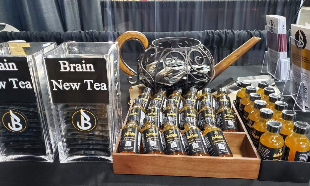 Display table featuring 'Brain New Tea' signs, with clear plastic stands and a wooden tray of small black bottles with gold caps labeled Brain New Tea Focus Shot.