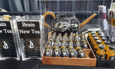 Display table featuring 'Brain New Tea' signs, with clear plastic stands and a wooden tray of small black bottles with gold caps labeled Brain New Tea Focus Shot.