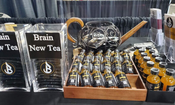 Display table featuring 'Brain New Tea' signs, with clear plastic stands and a wooden tray of small black bottles with gold caps labeled Brain New Tea Focus Shot.