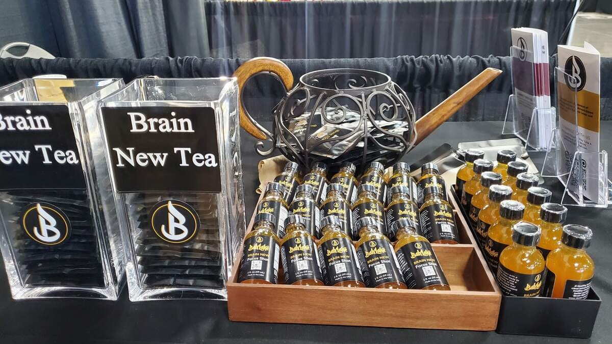 Display table featuring 'Brain New Tea' signs, with clear plastic stands and a wooden tray of small black bottles with gold caps labeled Brain New Tea Focus Shot.