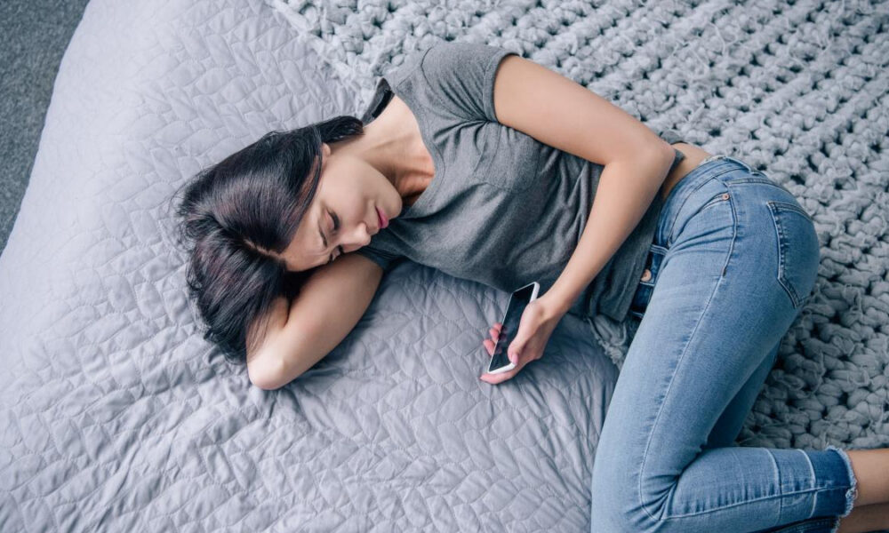 Woman sleeping on a gray quilted bed while holding a smartphone in one hand