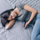 Woman sleeping on a gray quilted bed while holding a smartphone in one hand