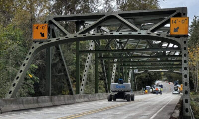 Green metal truss bridge under construction with 14'0" clearance signs and a Genie lift on the roadway, workers in the distance.