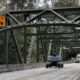 Green metal truss bridge under construction with 14'0" clearance signs and a Genie lift on the roadway, workers in the distance.