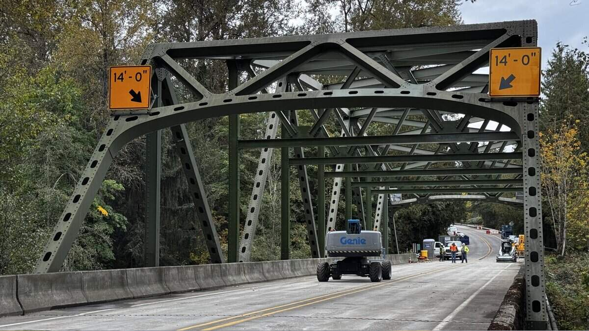 Green metal truss bridge under construction with 14'0" clearance signs and a Genie lift on the roadway, workers in the distance.