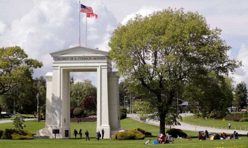 Wide park scene with a tall white arch monument labeled 'Children of a Common Mother', flags overhead, and people picnicking on the lawn.