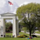 Wide park scene with a tall white arch monument labeled 'Children of a Common Mother', flags overhead, and people picnicking on the lawn.
