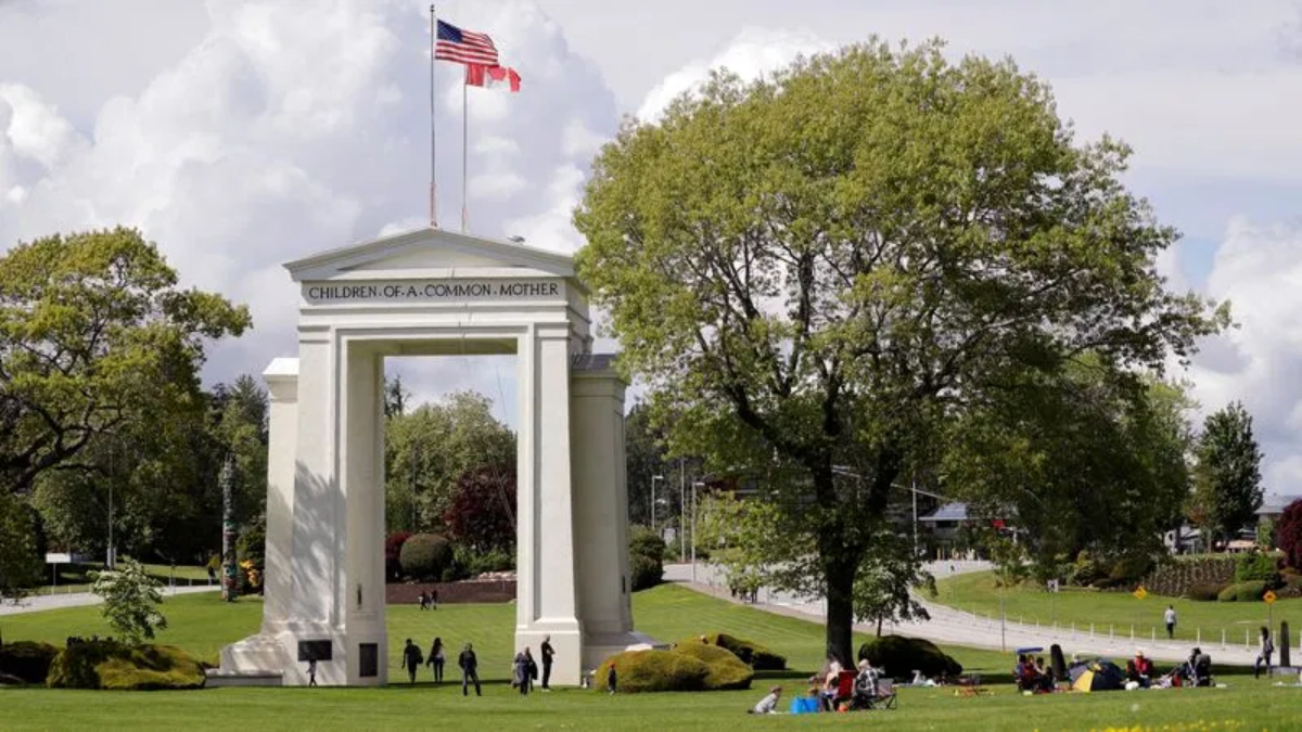Wide park scene with a tall white arch monument labeled 'Children of a Common Mother', flags overhead, and people picnicking on the lawn.