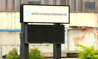 Outdoor sign for Nippon Dynawave Packaging Co. mounted on a dark metal frame in front of an industrial building.