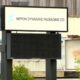 Outdoor sign for Nippon Dynawave Packaging Co. mounted on a dark metal frame in front of an industrial building.