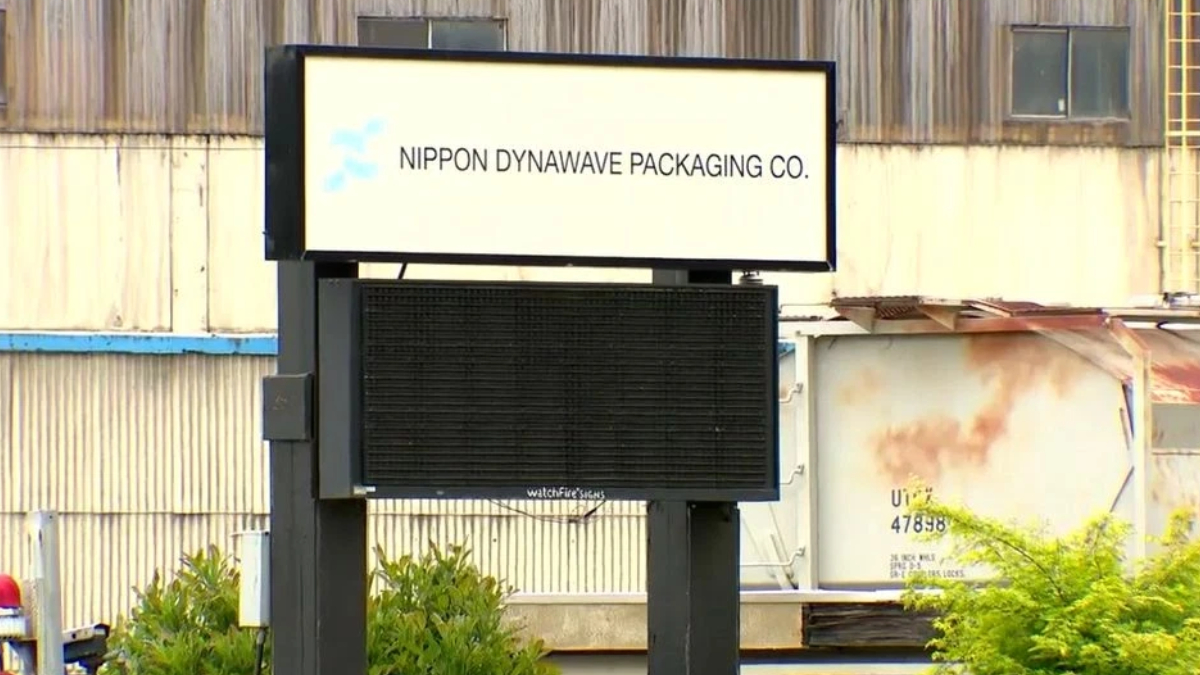 Outdoor sign for Nippon Dynawave Packaging Co. mounted on a dark metal frame in front of an industrial building.