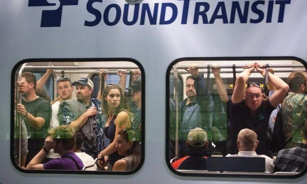 Crowded Sound Transit train car with passengers standing and seated, viewed through two windows.