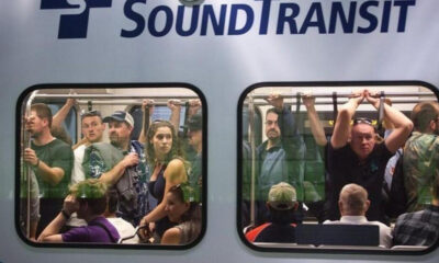 Crowded Sound Transit train car with passengers standing and seated, viewed through two windows.