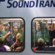 Crowded Sound Transit train car with passengers standing and seated, viewed through two windows.