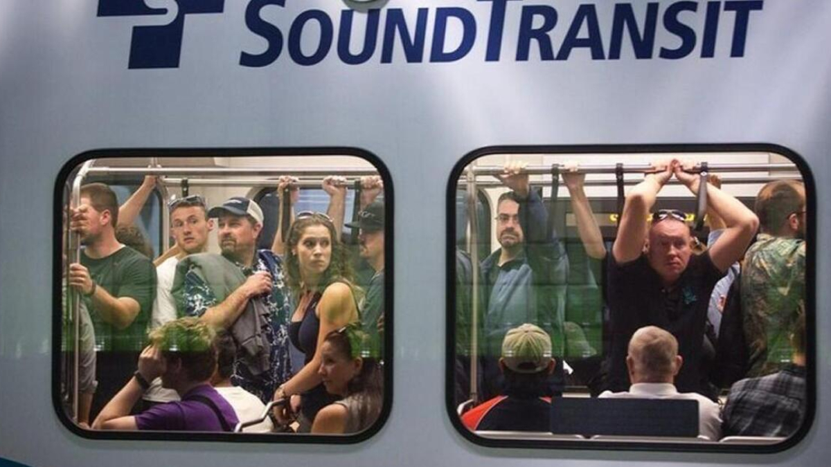 Crowded Sound Transit train car with passengers standing and seated, viewed through two windows.