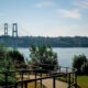 Suspension bridge spanning a river, viewed from a waterfront park with a curved railing and green bushes in the foreground.