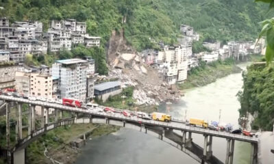 Bridge with loaded vehicles spanning a river, hillside town on both banks and a landslide debris pile on the slope nearby