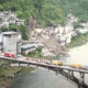 Bridge with loaded vehicles spanning a river, hillside town on both banks and a landslide debris pile on the slope nearby