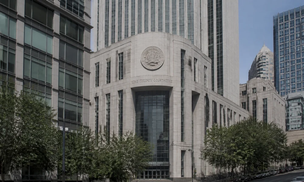 View of the King County Courthouse: a white stone building with a circular seal, flanked by tall glass skyscrapers, on a tree-lined street.