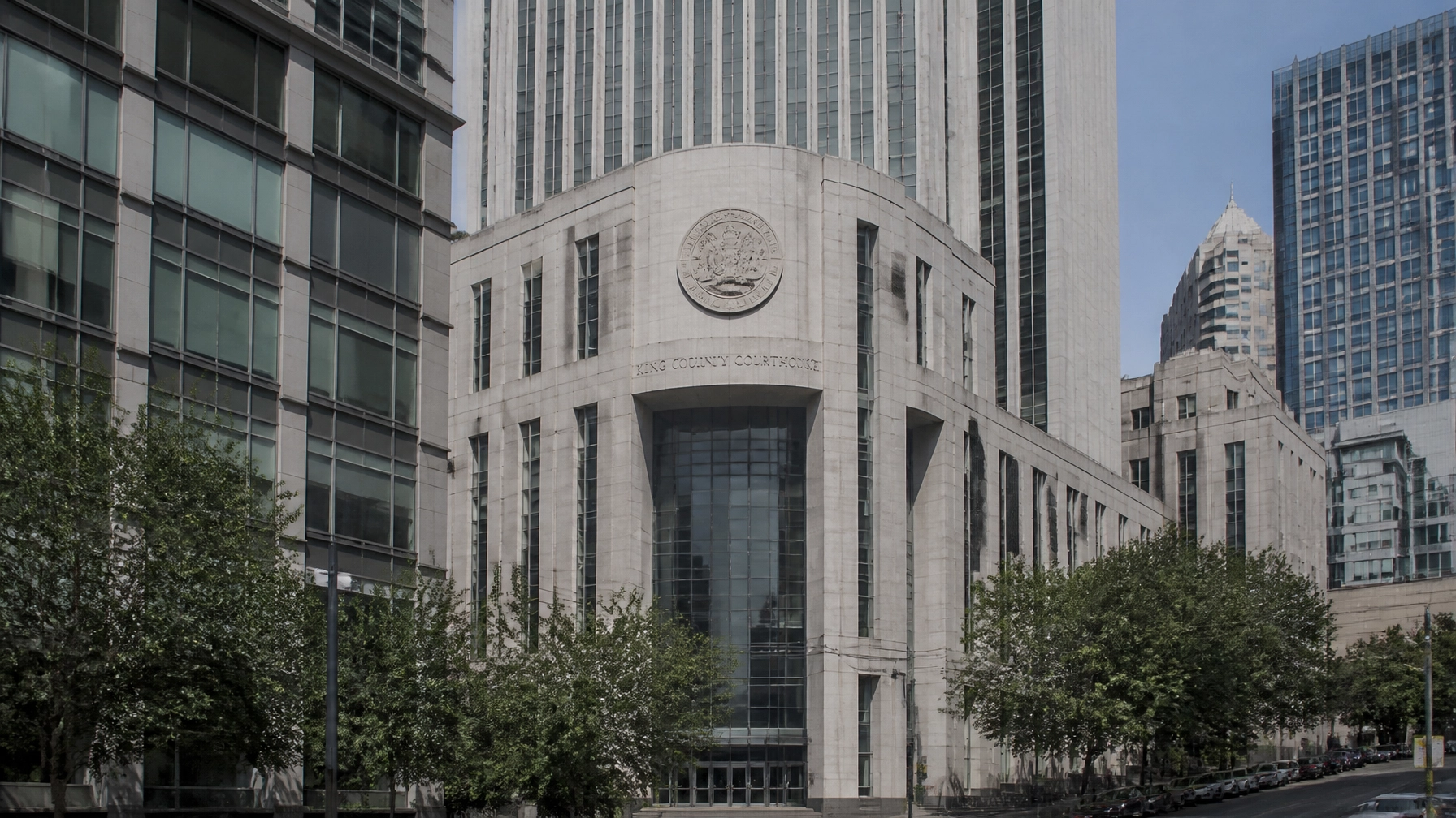 View of the King County Courthouse: a white stone building with a circular seal, flanked by tall glass skyscrapers, on a tree-lined street.