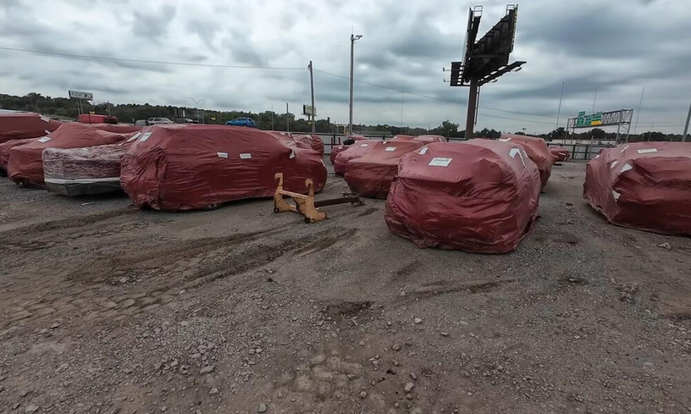 Red-wrapped vehicles or large objects lined up on a gravel lot under a cloudy sky, with a yellow tow hitch in the foreground