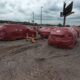 Red-wrapped vehicles or large objects lined up on a gravel lot under a cloudy sky, with a yellow tow hitch in the foreground