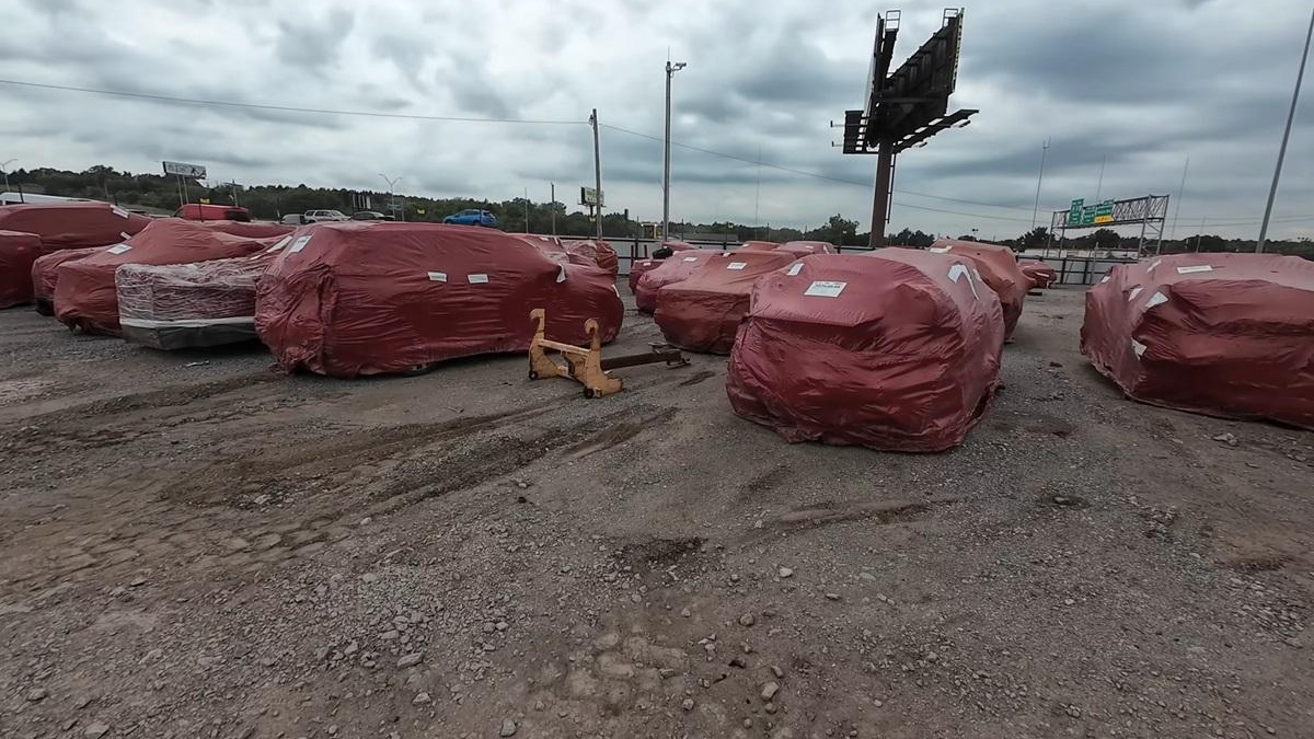 Red-wrapped vehicles or large objects lined up on a gravel lot under a cloudy sky, with a yellow tow hitch in the foreground