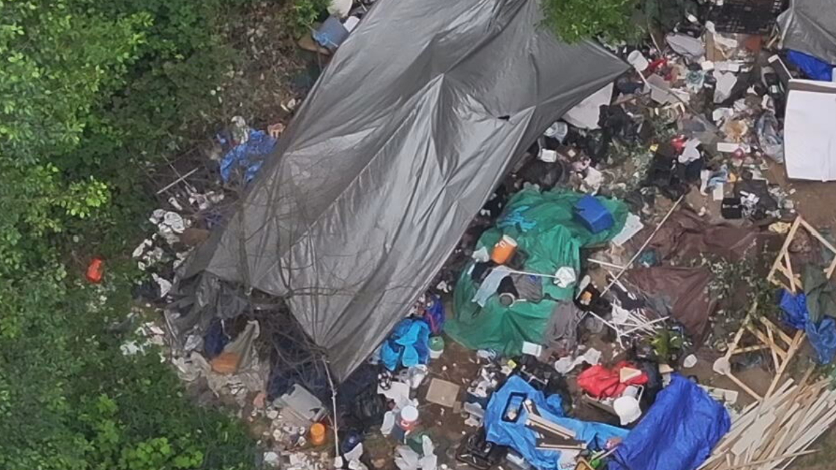 Aerial view of a cluttered encampment with tents, tarps, and piles of trash among trees.