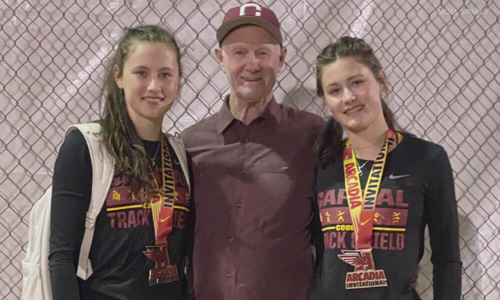 Two female track athletes wearing black uniforms and gold medals pose with a man in a maroon shirt and cap in front of a chain-link fence, at the Arkansas Invitational