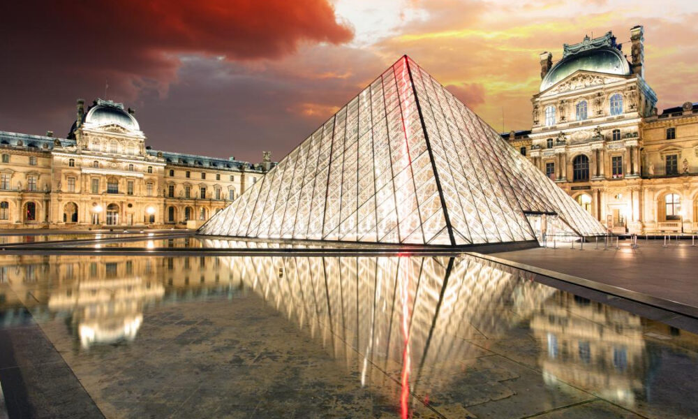 Glass pyramid lit at dusk in the Louvre Courtyard, with the historic building and reflecting pool nearby.