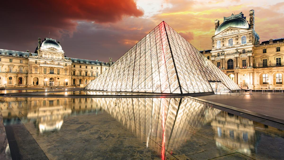 Glass pyramid lit at dusk in the Louvre Courtyard, with the historic building and reflecting pool nearby.