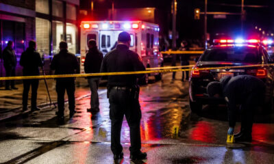 Nighttime street scene of a crime scene with police cars, an ambulance, yellow caution tape, and investigators on wet pavement behind the tape.