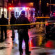 Nighttime street scene of a crime scene with police cars, an ambulance, yellow caution tape, and investigators on wet pavement behind the tape.