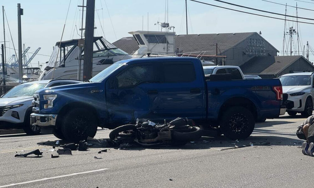 Blue Ford F-150 with front-end damage beside a downed motorcycle and debris on a harbor-side street.