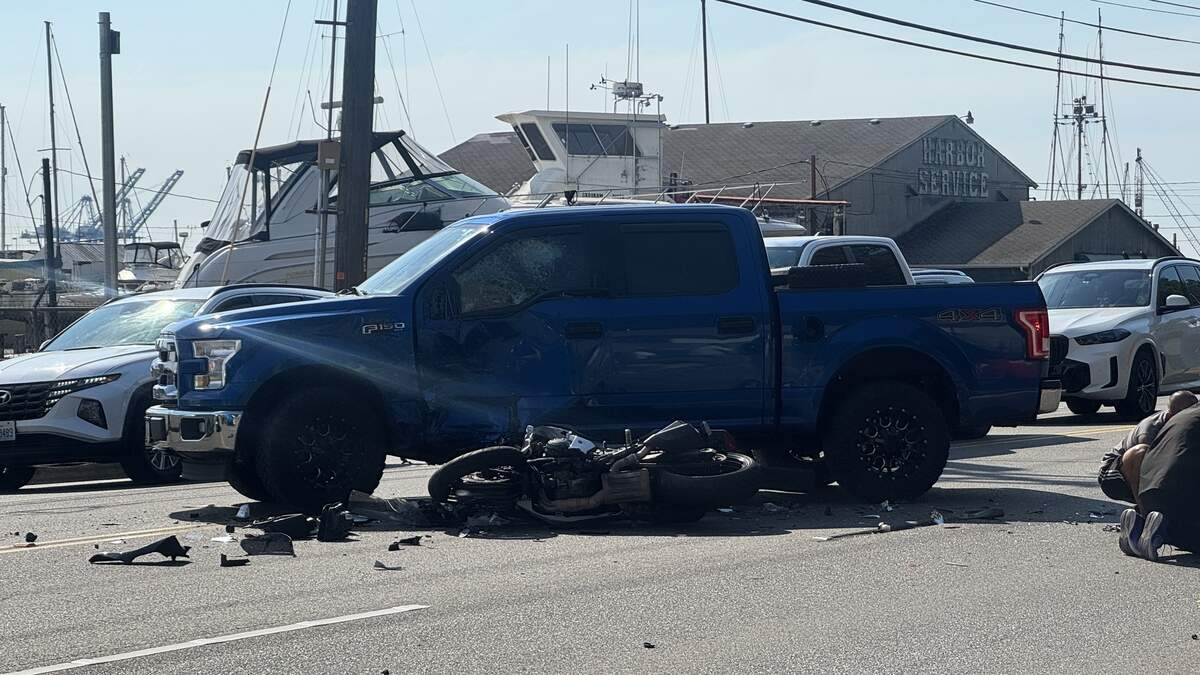 Blue Ford F-150 with front-end damage beside a downed motorcycle and debris on a harbor-side street.