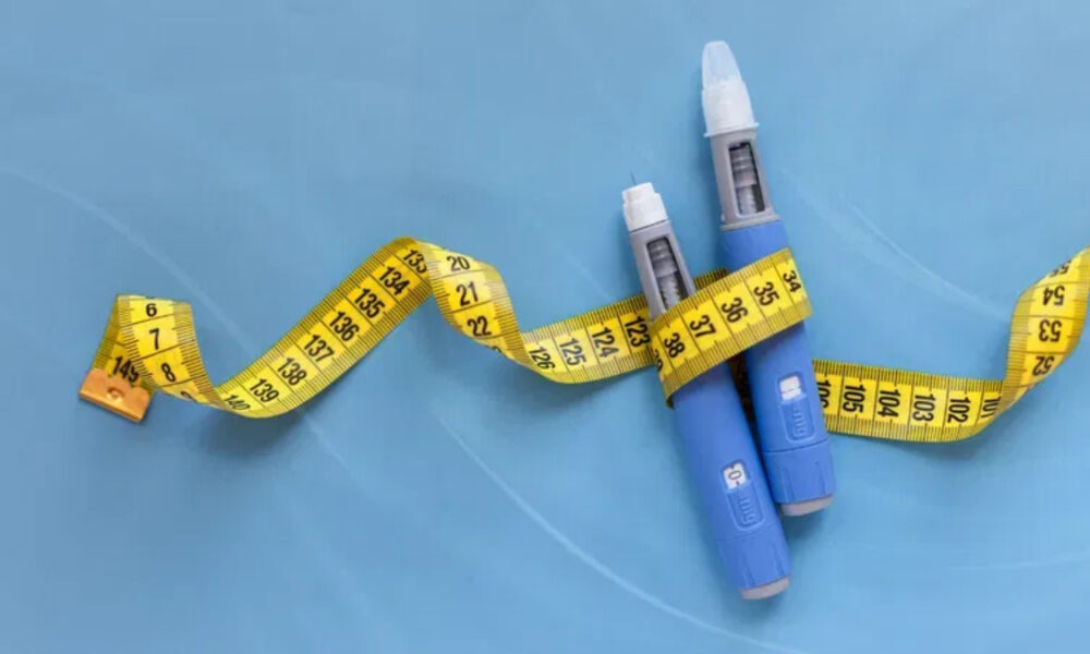 Two blue insulin pens with a yellow measuring tape draped around them on a blue background, symbolizing diabetes management.