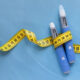 Two blue insulin pens with a yellow measuring tape draped around them on a blue background, symbolizing diabetes management.