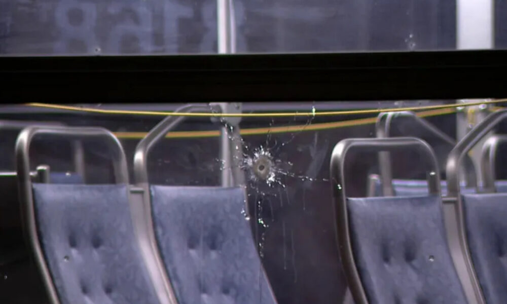 Bullet hole in a window with glass shatter and spray marks inside a vehicle with blue upholstered seats and metal frames.